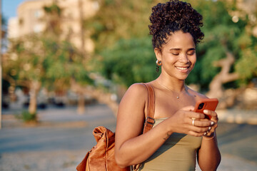 Young woman with curly hair and backpack using mobile phone while walking on a city street during summer holidays