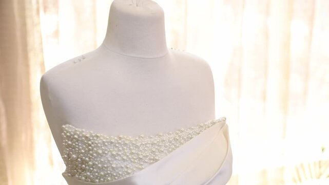 A stunning, voluminous white wedding gown is elegantly displayed on a mannequin. The strapless bodice features delicate pearl embellishments and a satin sash cinching the waist. 