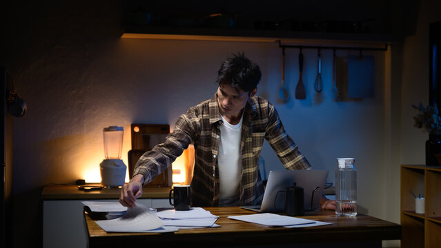 Focused man working late at night in kitchen, reviewing paperwork spread across table with laptop