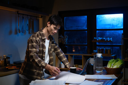 Young man working late night on laptop in a dimly lit kitchen, focusing on tasks with papers