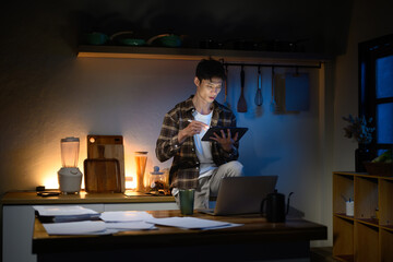 Asian man working late night in kitchen, reviewing data on digital tablet