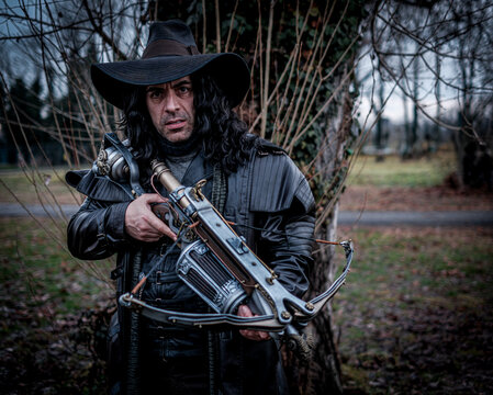 Milan, Italy 2025 01 26 Gabriel Van Helsing Cosplay A male figure in a black trench coat and wide-brimmed hat, wearing a mask, stands in the woods holding a futuristic, ornate steampunk crossbow.