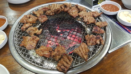 Korean barbecue pork grilling over hot charcoal on a wire mesh. Marinated meat pieces cooking with garlic and dipping sauce on the table. A popular and authentic Korean dining experience.
