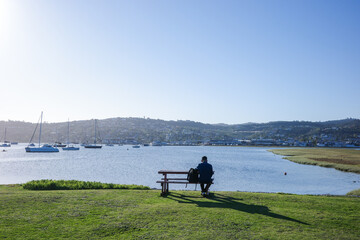 A peaceful landscape of a man sitting on a wooden bench, gazing at sailboats and yachts anchored in the calm bay. With lush green grass in the foreground and rolling hills in the background