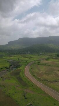 A drone shot follows a bike riding along the Nashik&ndash;Trimbak highway bordered by lush greenery and rolling fields. Above, the bright cloudy sky casts a gentle glow adding warmth to the scenic landscape