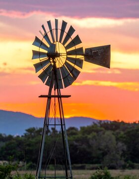 A vintage wind pump stands tall against a breathtaking sunset, silhouetted against vibrant colors of orange and pink, over a rolling landscape.