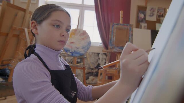 Portrait of creative young gilr wearing apron sitting in front of easel drawing picture with pencil during art master class