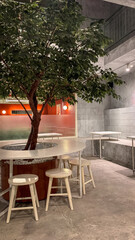 Modern cafe interior with a tree centerpiece, round table, and wooden stools, combining natural and minimalist industrial design.
