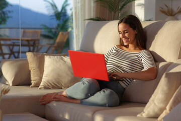 Happy woman at home at sunset using red laptop on a sofa