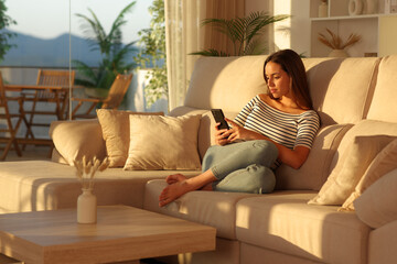 Obraz premium Relaxed woman at sunset at home using cell phone on a couch