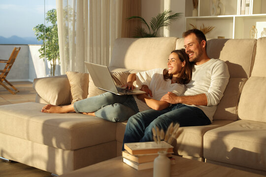 Happy couple at sunset using laptop sitting on a couch at home