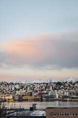A stunning panoramic view of a snow-covered coastal town in Norway, with mountains in the background and a body of water in the foreground. A soft, colorful sky adds a peaceful atmosphere.