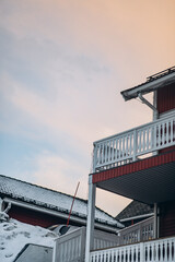 A low-angle view of traditional Scandinavian houses with snow-covered roofs and porches. The houses are painted red and feature white railings under a pastel-colored winter sky.