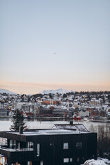 A wide-angle panorama of Troms&oslash;'s cityscape, featuring a dark modern house in the foreground, the fjord, and distant snow-covered mountains under a pale, wide sky with a tiny plane.
