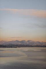 A serene winter landscape view of a fjord in northern Norway, with calm water reflecting the soft pastel sky. A large snow-covered mountain range dominates the background under a pale, wintry sky.