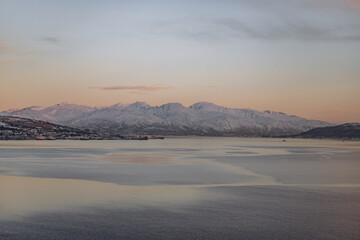 A wide-angle landscape of a tranquil fjord with snow-capped mountains on the horizon during a winter sunset. The calm water reflects the soft, pastel hues of the sky, creating a serene atmosphere