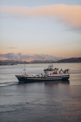 A large fishing boat crosses a calm fjord in Tromsø, Norway, with a stunning backdrop of snow-covered mountains under a soft, twilight sky, capturing the region's natural beauty and industry.