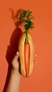 Naklejki Carrot hot dog in bread bun held by hand on orange background. Concept of vegan food, healthy lifestyle, creative snack design, vegetarian diet and wellness branding.