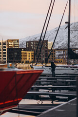 A person stands on a pier in a harbor, with sailing boats docked and modern buildings and a snow-covered mountain in the background. The scene blends urban life with natural beauty.