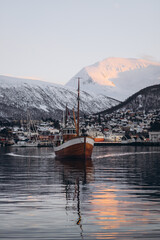 A classic wooden boat navigates the calm waters of the Tromsø fjord, with the city's buildings and majestic, snow-covered mountains illuminated by the setting sun in the background. © Tetiana