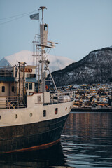 Naklejka premium A large boat is docked in the Tromsø harbor, with the city's twinkling lights and the snow-covered mountains providing a stunning backdrop during the calm winter evening.