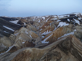 Aerial view of rugged, snow-dusted peaks carve deep valleys, a tapestry of ochre and charcoal under a pastel sky, The Highlands, Iceland.