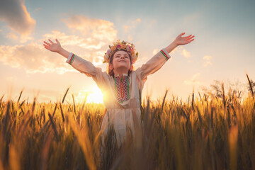 Beautiful young girl with flower chaplet, ethnic folklore dress with traditional Bulgarian embroidery during sunset on a wheat agricultural field