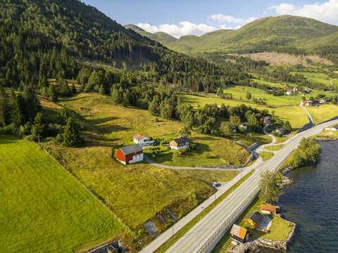 Aerial view of emerald green fields meet the serene waters of Jolstervatn, with a winding road connecting quaint red-roofed houses nestled against verdant hills, Jolstervatn, Sunnfjord, Norway.