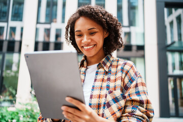 A joyful woman smiles brightly while using a tablet outdoors in a vibrant urban setting
