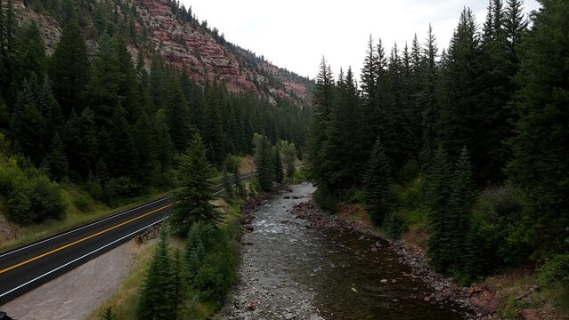 Aerial view of the Crystal River flowing alongside a road, with red rock cliffs and dense evergreen forests creating a striking contrast, Redstone, Colorado, United States.