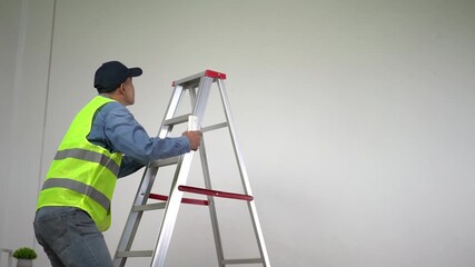 Construction worker wearing a safety vest and cap walking towards an aluminum ladder in a spacious, empty room with white walls, ready for home renovation work