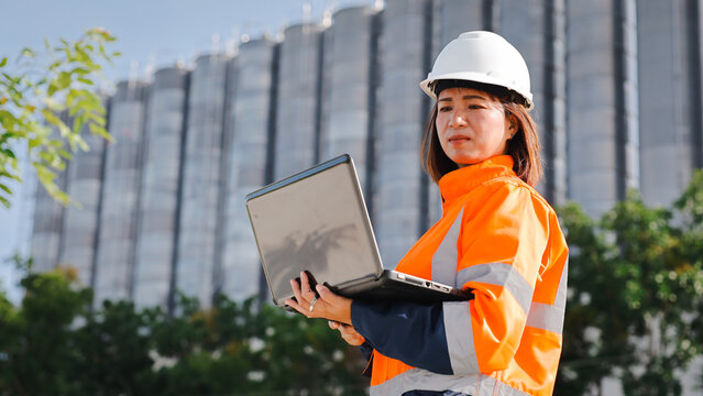 A construction worker in an orange safety vest and helmet stands outdoors, using a laptop while overseeing construction activities at a modern building site on a sunny day - Powered by Adobe