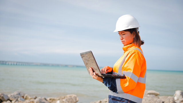 A construction engineer wearing a safety helmet and high-visibility jacket is focused on a laptop at a coastal site. The serene ocean backdrop enhances the professional atmosphere of the moment