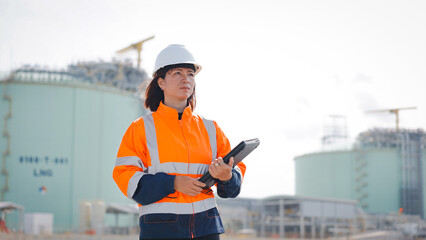 A safety-oriented professional observes operations at an LNG facility. She wears a helmet and an orange high-visibility jacket, holding a tablet in a busy industrial setting