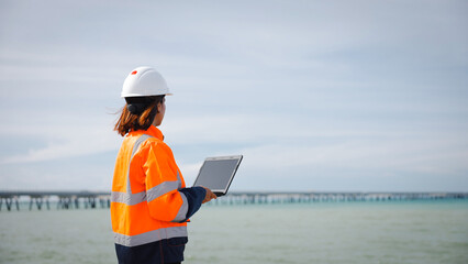 A safety-conscious engineer stands at the edge of the water, checking project details on a tablet....
