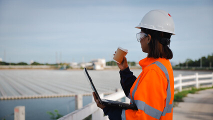 A woman in safety gear stands near water, holding a coffee cup in one hand and using a laptop in the other. She appears focused on her work at a construction site during the day