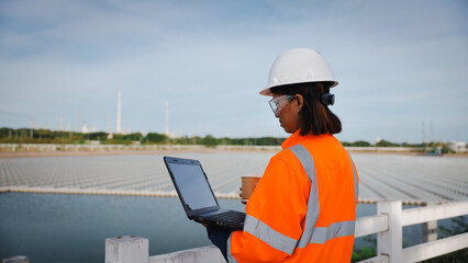 A professional wearing a hard hat and safety goggles stands by a water reservoir, using a laptop while holding a cup, overseeing a construction project during the day