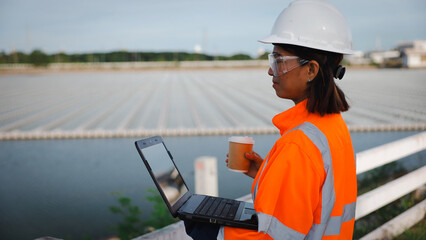 A female worker in an orange safety jacket and hard hat examines data on her laptop at an aquaculture facility near a waterway, holding a coffee cup, during daylight
