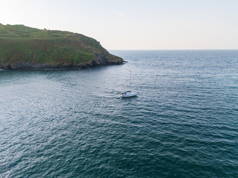 Aerial view of a lone sailboat gliding through the shimmering turquoise waters near a lush green headland, creating a striking contrast against the clear sky, Fowey, Cornwall, United Kingdom.