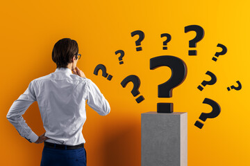 Businessman thinking while looking at floating question marks symbolizing problem solving and curiosity on orange background concept.