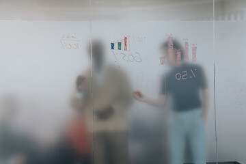 Colleagues brainstorm on a project, writing data and charts on a glass board during a business meeting. The blurred figures suggest a collaborative and dynamic work environment.