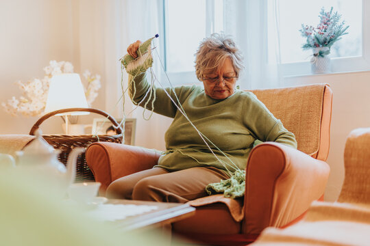 Senior woman sitting on an armchair knitting, surrounded by a cozy home interior with natural light and warm decor, reflecting a peaceful and fulfilling pastime of craft and serenity. - Powered by Adobe