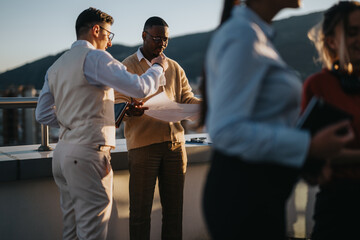 Multiracial business colleagues engage in a brainstorming session on a high-rise balcony at sunset. The collaborative environment reflects multicultural and diverse workforce dynamics in a modern