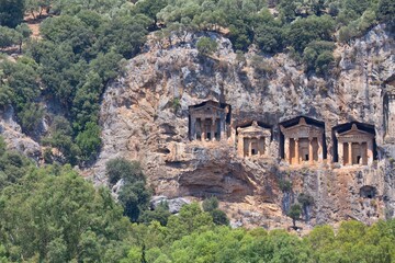 Fototapeta premium elaborately carved Lycian tombs are set into the face of a cliff, surrounded by green foliage. The cliff face appears to be a light tan or grey rock - Caunos Tombs of the Kings - Dalyan - Mugla 