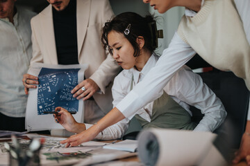 A diverse group of business coworkers collaborates on an architectural project in a bustling office environment. Blueprints and documents are spread across the table as they engage in team discussion.