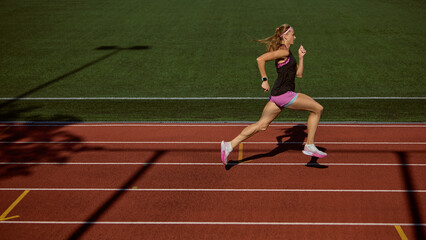 Fototapeta premium Sporty woman running on an open-air track under bright summer sunlight with determination. Concept of peak speed, physical effort, focus on victory, motion intensity, and athletic endurance.