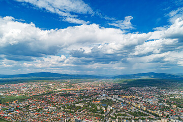 Fototapeta premium An aerial panoramic view of a city surrounded by mountains and greenery under a dramatic cloudy sky.