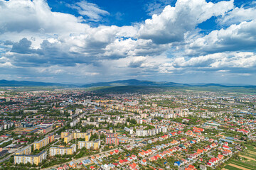 An aerial panoramic view of a city surrounded by mountains and greenery under a dramatic cloudy sky.