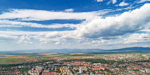 An aerial panoramic view of a city surrounded by mountains and greenery under a dramatic cloudy sky.