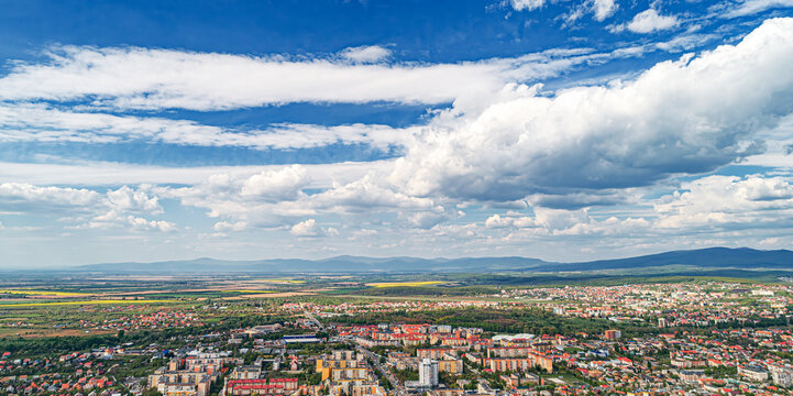 An aerial panoramic view of a city surrounded by mountains and greenery under a dramatic cloudy sky. - Powered by Adobe